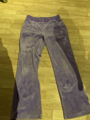 Lila velour mjukisbyxor Juicy Couture - Säljer ett par lila mjukisbyxor från Juicy Couture i mjuk velour. Byxorna har bred resår i midjan, raka ben och stora fickor framtill. Perfekta för en chill och avslappnad stil.
