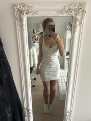 Vit kort klänning med snörning - Säljer en vit kort klänning från H&M med smala axelband och snörning framtill. Klänningen har ett broderat mönster och är rynkad längs hela kroppen för en snygg passform. Perfekt för sommarens alla tillfällen!