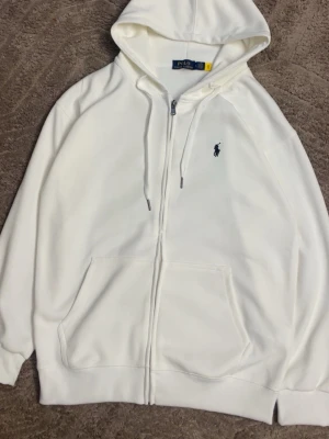 Vit hoodie från Polo Ralph Lauren - Snygg vit hoodie från Polo Ralph Lauren med dragkedja, huva med dragsko och klassisk blå logga broderad på bröstet. Hoodien har känguruficka framtill och ribbade muddar vid ärmar och nederkant. Perfekt för en clean och stilren look.
