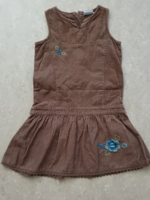 Klänning - Brun klänning i manchester med ärmlös design och broderade blå blommor på bröstet och kjolen. Klänningen har en volang nedtill och en enkel, avslappnad passform. Perfekt för dig som gillar unika detaljer och mjuka material.