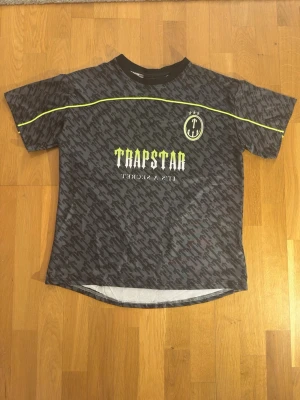 Trapstar svart mönstrad t-shirt - Snygg svart t-shirt från Trapstar med all-over mönster, neongula detaljer och tryck både fram och bak. På bröstet står det 'Trapstar' och på ryggen finns ett stort '22'-tryck. T-shirten har rund hals och korta ärmar, perfekt för en streetwear-look.