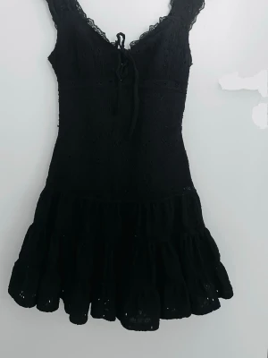 Svart spetsklänning med snörning🖤 - Svart kortklänning med spetsdetaljer och snörning framtill. Klänningen har volang nertill och är ärmlös med v-ringad hals. Perfekt för dig som vill ha en feminin och trendig look.