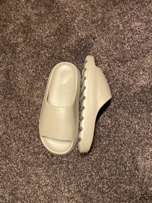 Beigea Yeezy slides - Säljer ett par beigea yeezy slide med öppen tå och chunky räfflad sula. Tofflorna är gjorda i ett mjukt och flexibelt material som känns skönt på foten. Perfekta för chill dagar hemma eller när du vill ha en clean och enkel look.