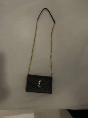 Svart YSL kuvertväska med kedja - Säljer en svart kuvertväska från Saint Laurent med guldfärgad kedjerem och ikoniskt YSL-emblem framtill. Väskan är tillverkad i präglat läder med snyggt chevronmönster. Perfekt accessoar för dig som gillar lyxig och stilren design.