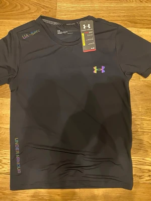 Svart träningstopp från Under Armour - Sportig svart topp från Under Armour med färgglad logga på bröstet och tryckt text på axeln och nederkanten. Tillverkad i lätt och snabbtorkande syntetmaterial, perfekt för träning. Kortärmad och med rund halsringning.