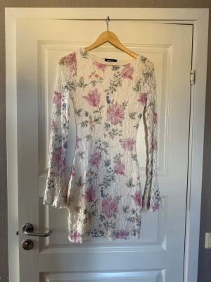Blommig vit spetsklänning - Söt kort klänning i vit spets med blommigt mönster i rosa, blått och grönt. Klänningen har långärmade ärmar med lätt utsvängda ärmslut och en figurnära passform. Perfekt för sommarens alla tillfällen.