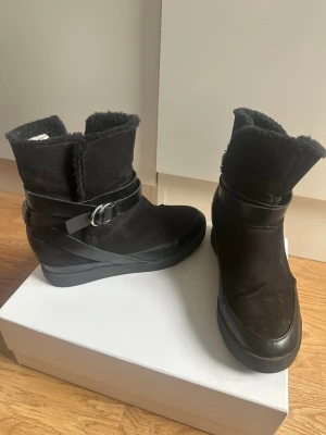Svarta kilklackade boots med fuskpäls - Snygga svarta boots med kilklack, fuskpälsfoder och dekorativa remmar med spänne runt ankeln. Ovandelen är i mockaimitation och sulan är i gummi. Perfekta för kalla dagar när du vill vara både varm och trendig.