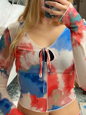 Färgstark mesh topp med knyt - Trendig mesh topp med lång ärm och färgstarkt abstrakt mönster i rött, blått och vitt. Toppen har v-ringning med knyt framtill och är croppad i modellen. 