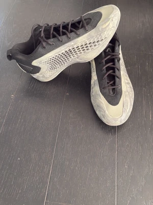 Adidas svartgrå sneakers med meshdetaljer - Säljer ett par svarta och grå Anthony Edwards basket skor med futuristisk design och meshdetaljer på sidorna. Ovandelen är i textil och syntet, med svarta skosnören och en unik mönstrad sula. Perfekta för dig som gillar basket och som tränar på fritiden.