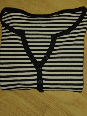 Randig långärmad Henley topp med knappar - Svartvit randig långärmad Henley topp med knappar. Från märket The set. Den kommer med 1 extra knapp. Den är gjord av  95% bomull och 5% elastan. Den är helt ny. Har aldrig använt den förutom när jag har provat den. Den är i storlek XS. Det står på deras hemsida XS (EU34). Den är gjord i Bangladesh. Jag kan sänka den om det behövs.