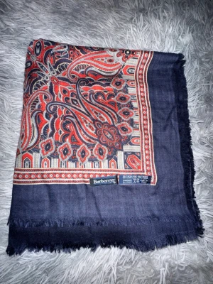 Mönstrad halsduk från Burberrys - Snygg halsduk från Burberrys i en mix av 65% ull och 35% siden. Mörkblå botten med paisleymönster i rött, beige och svart. Tunt tyg med fransade kanter och exklusiv känsla. Tillverkad i Italien och perfekt för att lyfta din outfit.