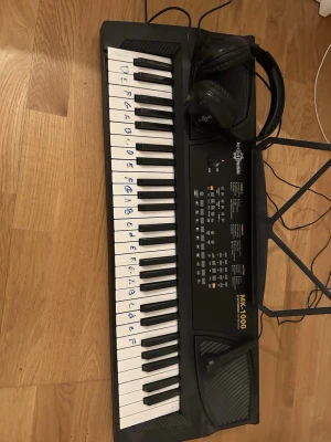 MK-1000 keyboard med mikrofon & hörlurar - Elektroniskt keyboard MK-1000 i svart plast med vita och svarta tangenter, där tonnamn är markerade på vissa tangenter. Medföljer mikrofon och svarta hörlurar. Perfekt för dig som vill spela och experimentera med olika ljud och funktioner.
