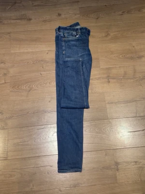 Jeans  - Säljer ett par klassiska blå jeans med rak passform och fem fickor. Jeansen har en snygg läderpatch bak i midjan och kontrastsömmar. Tillverkade i slitstark denim, perfekta för en avslappnad och tidlös stil.