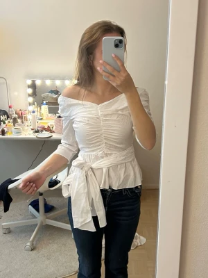 Vit offshoulder blus UTAN knytband - TRÖJAN KOMMER INTE MED KNYTBAND! Snygg vit blus med offshoulder-design och trekvartsärm. Blusen har rynkade detaljer framtill och ett brett knytband i midjan som ger en feminin siluett. Materialet ser ut att vara bomull eller liknande och toppen har en lätt peplum-effekt nedtill.