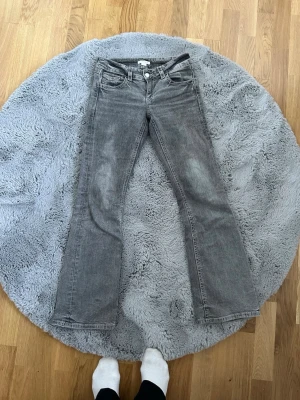 Grå bootcut jeans med slitningar - Säljer ett par grå bootcut jeans med lätt slitna detaljer och klassisk femficksdesign. Jeansen har en låg midja och raka ben som blir lite vidare nedtill. Perfekta för dig som gillar en avslappnad och cool stil.