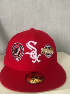 Röd Chicago White Sox keps New Era 7 1/4 - Snygg röd keps från New Era med Chicago White Sox-logga framtill, broderade World Series-detaljer och patchar på sidorna. Klassisk 59FIFTY-modell med platt skärm och broderade ventilationshål. Perfekt för dig som gillar sportig streetwear.