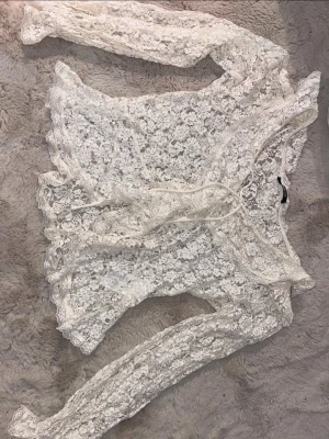 Vit spetsblus från Gina Tricot - Säljer en superfin vit spetsblus från Gina Tricot. Blusen har långa ärmar, omlottknäppning med knytband i midjan och är helt i genombruten spets med blommigt mönster. Perfekt för dig som gillar romantisk och trendig stil.