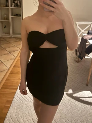 Svart axelbandslös cut out-klänning - Trendig svart axelbandslös miniklänning med twistad byst och cut out-detalj framtill. Klänningen har figurnära passform och är perfekt för dig som gillar en stilren men edgy look. Dragkedjan har gått sönder men går säkert att fixa enkelt hos en skräddare eller om man är duktig på att sy!!