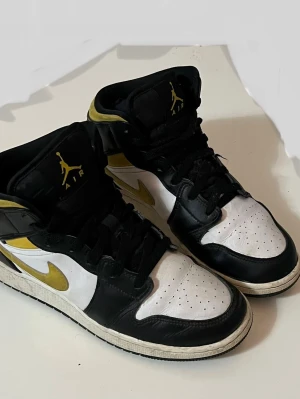 Nike Air Jordan 1 Mid svart/vit/guld - Säljer ett par Nike Air Jordan 1 Mid sneakers i svart, vitt och guld. Skorna har klassisk hög siluett, svart läder med vita paneler och guldfärgade detaljer på swoosh och logga. Vit sula och svarta skosnören. Perfekta för dig som gillar streetwear och ikonisk design. Hör av dig vid intresse. 🎉😆
