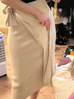 Beige ribbad omlottkjol med knyt - Trendig beige omlottkjol i ribbat material med knyt i midjan. Kjolen har en smickrande midilängd och sitter snyggt figurnära. Perfekt för dig som gillar stilrena och moderna plagg.