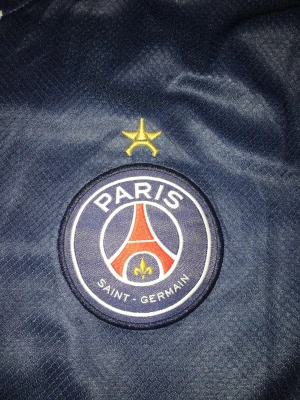PSG matchtröja i mörkblått från Nike - Säljer en mörkblå Paris Saint-Germain matchtröja från Nike med klubbmärke och gul stjärna på bröstet. Tröjan har vita och röda detaljer samt tryck på insidan av kragen. Perfekt för dig som älskar fotboll och vill stötta PSG. Fraktar inom 24 timmar