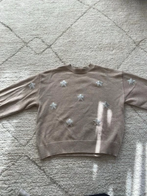 Beige stickad tröja med stjärnor - Beige stickad tröja med rund hals och långa ärmar från Gina Tricot. Storlek 146/152. Tröjan har silvriga paljett stjärnor framtill som ger en söt och trendig look. Ribbstickade muddar vid ärmslut och nederkant. Perfekt för dig som gillar en stilren men lekfull stil.