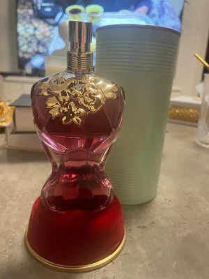 Jean Paul Gaultier parfymflaska - Säljer en ikonisk parfym från Jean Paul Gaultier i en djup röd glasflaska formad som en kvinnokropp. Flaskan har guldfärgade blommor upptill och står på en rund röd sammetsbas med guldkant. Perfekt för dig som gillar lyxiga och unika dofter.