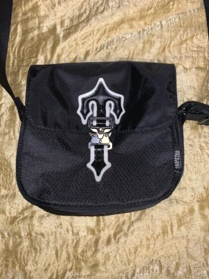 Svart axelväska från Trapstar - Säljer en svart axelväska från Trapstar med justerbar rem och coolt spänne framtill. Väskan har en unik vit och svart detalj på locket och är tillverkad i slitstarkt tyg. Perfekt för dig som vill ha en streetwear-inspirerad look.