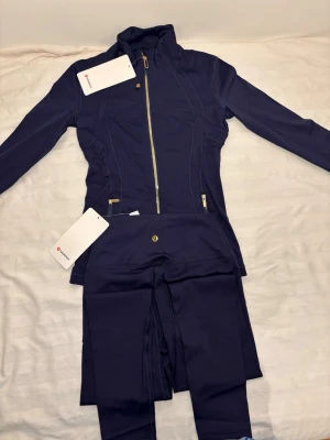 Mörkblå träningsset från Lululemon - Snyggt mörkblått träningsset från Lululemon med figursydd jacka och matchande tights. Jackan har dragkedja framtill, höga fickor med dragkedja och ståkrage. Materialet är stretchigt och följsamt, perfekt för träning eller chill. (Storlek 4) vi kan diskutera pris 