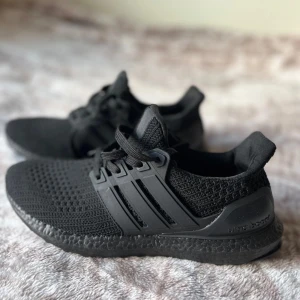 Svarta Adidas Ultraboost sneakers - Säljer ett par helsvarta Adidas Ultraboost sneakers med stickad ovandel och klassiska tre ränder på sidan. Skorna har snörning, platt sula och en riktigt skön boost-dämpning. Perfekta för dig som gillar stilrena och bekväma sneakers till vardags eller träning. HELT NYA-ALDRIG ANVÄNDA