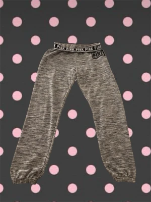 Grå mjukisbyxor från PINK - Säljer ett par gråmelerade mjukisbyxor från PINK med bred resår i midjan där det står 'PINK' upprepat. Byxorna har muddar vid bensluten och tryckt logga längs ena benet. Perfekta för chill dagar och riktigt sköna att ha på sig.