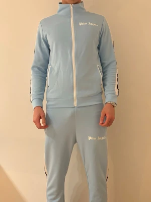 Ljusblå sett från Palm Angels - Snyggt ljusblått träningsset från Palm Angels med vita och svarta ränder längs ärmar och ben. Setet består av en zip-jacka med hög krage och matchande byxor, båda med logga tryckt i vitt. Perfekt för sport eller chill. Materialet är mjukt och stretchigt.