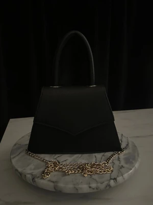 Svart classy handväska  - Stilren svart handväska i slät läderimitation med topphandtag och elegant guldfärgad kedja. Väskan har en strukturerad, trekantig form som ger en modern vibe.