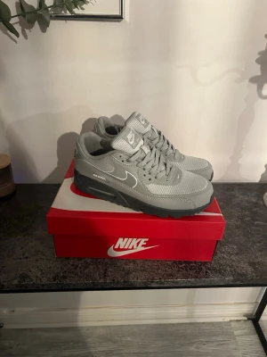 Nike Air Max 90 grå sneakers - Snygga Nike Air Max 90 sneakers i helgrå färg med vit swoosh och detaljer. Skorna har en klassisk siluett, mesh och syntetmaterial på ovandelen samt den ikoniska Air Max-sulan för extra komfort. Perfekta för dig som gillar stilrena och sportiga sneakers.