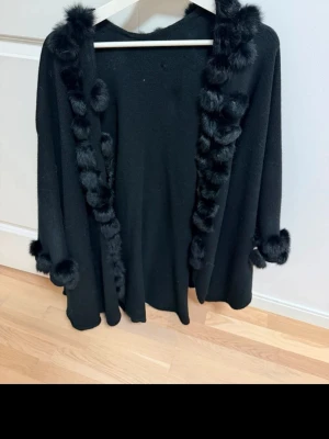 Svart kappa med fuskpälsdetaljer - Stilren svart kappa med fluffiga fuskpälsdetaljer längs krage, framkant och ärmslut. Kappan har en rak och lös passform som ger en trendig look. Perfekt för dig som vill ha en elegant och mysig ytterjacka med det lilla extra. Poncho