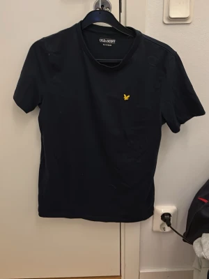Svart t-shirt från Lyle & Scott - Klassisk svart t-shirt från Lyle & Scott med gul broderad logga på bröstet. T-shirten har rund halsringning och korta ärmar. Tillverkad i mjuk bomull för en skön känsla och enkel stil.