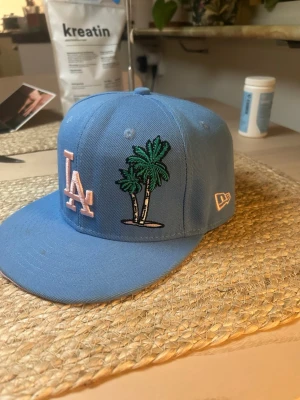 Blå LA keps med palmer från New Era - Snygg blå keps från New Era med LA-logga i ljusrosa framtill och broderade palmer. Kepsen har platt skärm och broderad jubileumspatch på sidan. Perfekt för dig som gillar streetstyle och sportiga accessoarer.