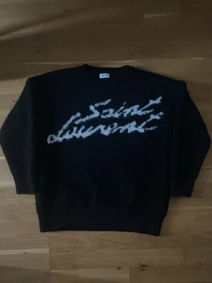 Svart stickad Saint Laurent-tröja - (PRISEN KAN VERKLIGEN DISKUTERAS)Svart stickad tröja från Saint Laurent med vit, suddig logotyp framtill. Oversized passform och rund halsringning. Perfekt för dig som gillar streetstyle och exklusiva märken.