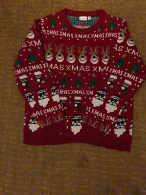 Röd jultröja med tomtar och renar - Säljer en röd stickad jultröja med mönster av tomtar, renar, julgranar och texten 'XMAS' i vitt. Tröjan har rund hals och långa ärmar. Perfekt för dig som vill maxa julkänslan med en färgglad och lekfull design.