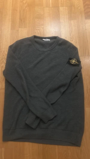 Grå sweatshirt från Stone Island - Säljer en stilren grå sweatshirt från Stone Island med den klassiska loggan på vänster ärm. Tröjan har rund halsringning och ribbade muddar vid ärmslut och nederkant. Perfekt för dig som gillar streetwear och vill ha något enkelt men snyggt.