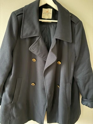 Mörkblå trenchcoat Holly & Whyte - Stilren mörkblå trenchcoat från Holly & Whyte by Lindex med dubbelknäppning och guldknappar. Klassisk krage och två snedställda fickor framtill. Fodrad insida för extra komfort. Perfekt för dig som gillar tidlös och elegant stil.