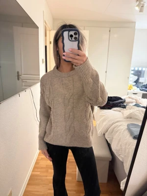 Beige kabelstickad tröja - En beige kabelstickad tröja med rund hals och ribbade muddar. Tröjan har ett klassiskt mönster framtill och är lite oversized med långa ärmar. Perfekt för att hålla sig varm under kyliga dagar.