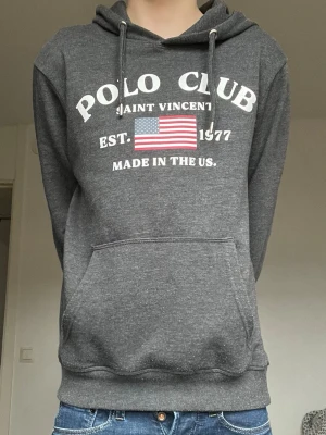 Mörkgrå hoodie Polo Club - Mörkgrå hoodie från Polo Club Saint Vincent med vit text och amerikansk flagga på bröstet. Klassisk känguruficka framtill och justerbar huva med snören. Perfekt för en avslappnad stil.