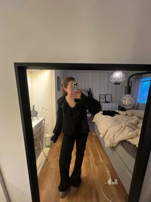 Svart blus med volang och v-ringning - Säljer en svart blus med v-ringning och stora volangdetaljer runt halsen. Blusen har långa ärmar och en loose passform som ger en avslappnad och trendig look. 