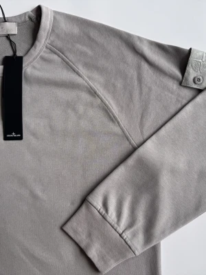 Stone Island ghost  - Snygg grå sweatshirt från Stone Island med klassisk rund hals och ribbade muddar. Tröjan har den ikoniska Stone Island-loggan på ärmen och är tillverkad i mjuk bomull för maximal komfort.