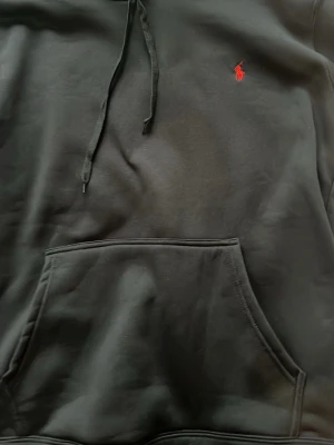 Svart hoodie från Ralph Lauren - Säljer en svart hoodie från Ralph Lauren med klassisk huva och dragsnören. Tröjan har en röd broderad logotyp på bröstet och en stor magficka. Materialet är mjukt och bekvämt, perfekt för en avslappnad stil. Fick som present har inte kommit till nån användning.passar även m den är stor i storlek 