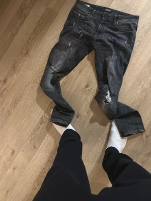 Mörkgrå slitna jeans från Nudie - Säljer ett par mörkgrå jeans från Nudie med slitningar och hål på knäna. Jeansen har klassisk femficksmodell och normal passform. Perfekta för dig som gillar en avslappnad och trendig stil.