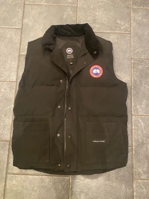 Svart dunväst från Canada Goose - Stilren svart dunväst från Canada Goose med klassisk logga på bröstet och två stora fickor framtill. Västen har hög krage, tryckknappar och dragkedja. Perfekt för lager-på-lager och håller dig varm under kyliga dagar.
