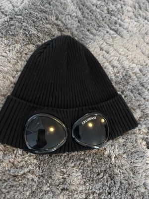 Svart mössa med glasögondetalj C.P. Company - Säljer en svart ribbstickad mössa från C.P. Company med ikoniska glasögonlinser framtill. Mössan är gjord i mjukt material och har en uppvikt kant. Perfekt för dig som vill sticka ut med en unik accessoar.