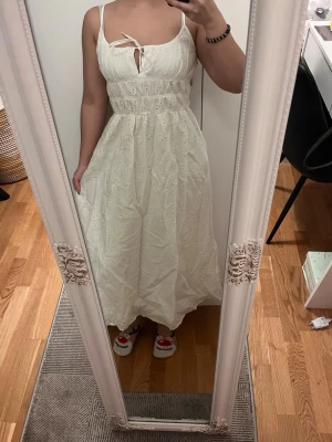 Vit långklänning med broderade detaljer - Superfin vit långklänning med smala axelband och broderade detaljer. Klänningen har en söt knytning framtill och är gjord i ett lätt bomullstyg, perfekt för varma dagar. Den har en feminin passform och faller snyggt längs kroppen.